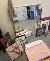 Photo showing large gold rectangular mirror leaning against wall with four framed art pieces on the floor nearby and macrame plant hanger hanging on wall.