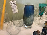 Wide view showing assortment of glassware: tall blue art glass vase, clear pitchers, yellow glass candlestick, green painted wine glass, small glass bowl.