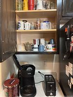 Shelf with various mugs, glasses, and bottles above coffee pot and toaster on counter
