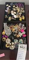 Full view of assorted vintage and modern costume jewelry on a black tray including earrings, rings, and pins.