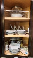 Metal shelf inside wooden cabinet with stacks of white dinnerware plates, bowls, mugs, and deviled egg plate at bottom right.