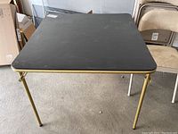 Black folding card table with gold-colored metal frame and surface showing some scuffs and wear, sitting on concrete floor in garage.