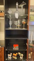 Full view of cabinet showing glassware on upper shelves, painted horse figurines on lower shelves, and a small box with various items.