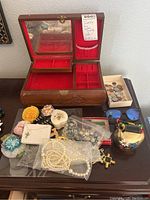 Photo showing open Asian style wooden jewelry box with red felt lining, various jewelry pieces including flower brooches, pearl necklace, pins, earrings, miniature cowbell, and a box of coins alongside blue sunglasses.