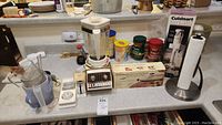 Photo showing vintage Osterizer blender, Cuisinart hand blender in box, Sunbeam electric carving knife in box, spices, kitchen timer, glass food processor bowl, metal towel paper holder with paper towel roll on counter.