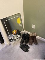 Photo showing a rectangular mirror with white and yellow floral decorative art, accompanied by three pairs of women's shoes: black lace-up boots, brown fur-lined boots, and black strappy sandals.