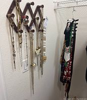Photo showing wooden accordion-style necklace holder with multiple gold-tone necklaces with different pendants hanging on wall. To the right side, men's ties in various colors and patterns hang on a tie rack. Mardi Gras bead necklaces are partially visible behind the door.