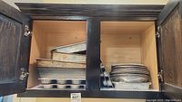 Open dark wooden kitchen cabinet showing stacked assortment of metal baking pans including muffin tins, sheet pans, loaf pans, and a round pizza pan.