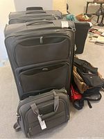 Photo showing two large and medium gray Samsonite wheeled suitcases in soft fabric with front zippered pockets, multiple backpacks and bags in black, brown, red fabric stacked around them