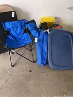 Photo of one blue folding camp chair and camping chair bags beside two large plastic storage tubs with lids and cardboard boxes in a garage setting