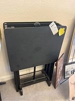 Tray tables folded and stacked on the stand. Tables and stand are black with flat rectangular trays and metal legs.