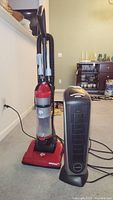 Photo of Dirt Devil red and black upright vacuum cleaner next to black Lasko ceramic heater on carpeted floor in living room.