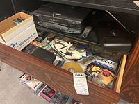 Drawer containing various cassette tapes, CDs, DVDs, a VHS VCR player, remote controls, cables, and other media accessories