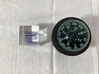 A rectangular aluminum piece encased in clear block with blue printed text about the 1928 Ford Trio Tour and first South Pole flight next to a black-cased aviation compass with green luminous markings.