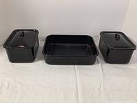 Front view of two black enamelware rectangular lidded bowls on either side of a black rectangular enamelware tray on white fabric background.