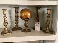 Four brass candle sticks with gold glitter glass orb on brass stand displayed on white shelf.