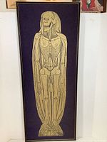 Full view of framed brass rubbing on purple fabric showing skeleton figure with long hair and detailed bones.