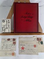 Photo of red Modern Postage Stamp Album with vintage postal covers in front, some worn and with visible stamps and cancellation marks