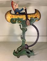 Full view of metal art whirligig showing Native American wearing headdress rowing a yellow canoe with green waves base.