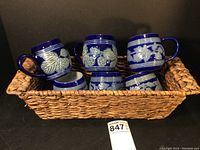 Photo showing six royal blue stoneware mugs with white strawberry and leaf relief designs sitting inside a rectangular woven basket.