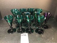 Front view of all 12 cordial glasses grouped together, showing 11 green glasses and one purple glass all hand-blown with black stems.