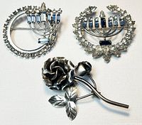 Top view of all three silver brooches on white background: one rose brooch and two crystal jeweled menorah brooches