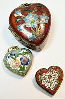 Front view of three heart-shaped cloisonné enamel jewelry items showing a locket pendant, a smaller pendant, and a brooch with floral designs