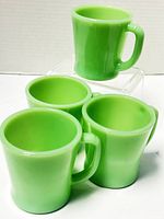 Four Fire-King jadeite green mugs, one raised above others showing shape and color.