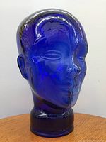 Front three-quarter view of cobalt blue glass head sculpture showing stylized facial features and cylindrical pedestal