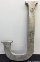 Front view of the large cast aluminum letter J showing its height, thickness, and patina on silver-gray metal.
