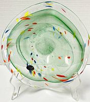 Top-front angled view of a Murano blown glass bowl with green tint and multicolored flecks shown on a clear display stand.