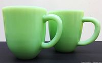 Two light green Jadite glass mugs side by side showing front angled view with handles visible.