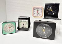 Full set of five travelling alarm clocks arranged on a white surface showing varied vintage designs and colors.