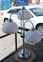 Full view of the 3 shade tri-light table lamp with chrome base and white shades, shown outdoors with background car and storage containers.