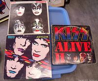Front covers of 3 KISS vinyl LP albums: Dynasty, Asylum, and Alive II placed on blue surface.