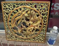 Square carved wood fretwork screen showing dragon and phoenix motif with polychrome and gold patina, photographed outdoors on a brick surface next to a water bottle for scale.
