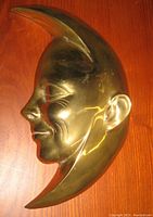 Brightly lit photo showing the side profile of the gilt metal moon face sculpture, placed on a wood surface.