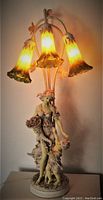 Front view of illuminated lady and child table lamp with 3 bell-shaped art glass shades resembling flowers, showing detailed sculptural base and warm light effect.