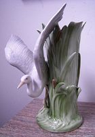 Front angle of crane bird vase showing outstretched wing, detailed feathers, and green grass base with cattails.