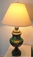 Full view of the table lamp lit, showing beige lampshade and ornate green and gold glass base with metal accents.