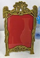 Front view of rectangular ornate brass frame showing decorative top with leaf motifs and red backing