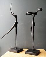 Pair of bronze abstract male and female statues on square bases shown against gray wall with natural light.