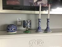 Four candle holders displayed: two tall blue and white taper candle holders with white candles, blue rounded votive candle holder with star and circle cutouts, and another blue patterned votive holder on green base.