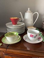 Photo of unmatched and matched tea cups, saucers, plates, and white coffee pot with gold trim arranged on a wooden surface