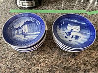 Two stacks of blue and white round collector plates laid on a granite countertop showing detailed painted scenes dated with years like 1973 and 1960.