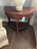 Front view of demi lune wooden side table with lower shelf, lamp placed on top but not included in lot.