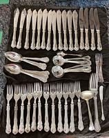 Flatware spread on dark cloth including knives, spoons, forks, and serving pieces showing ornate handles and polished surfaces