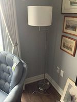 Floor lamp standing next to a blue chair near a window, showing full height and shade.