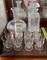 Glass and crystal decanters and 10 small stemmed glasses arranged on a wooden surface with a mirror background reflecting the pieces.