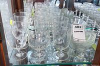 Wide view showing a large set of clear stemmed wine glasses and bowls with two etched designs, wheat and fluted, arranged on glass shelves.